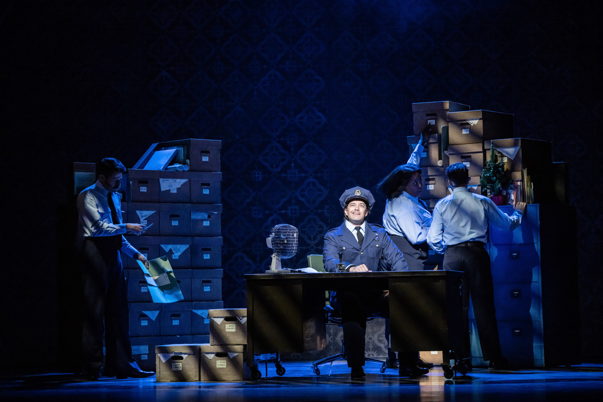 Lee Mead sitting at a desk in a police uniform as 'Eddie Souther' in 'Sister Act' on the West End