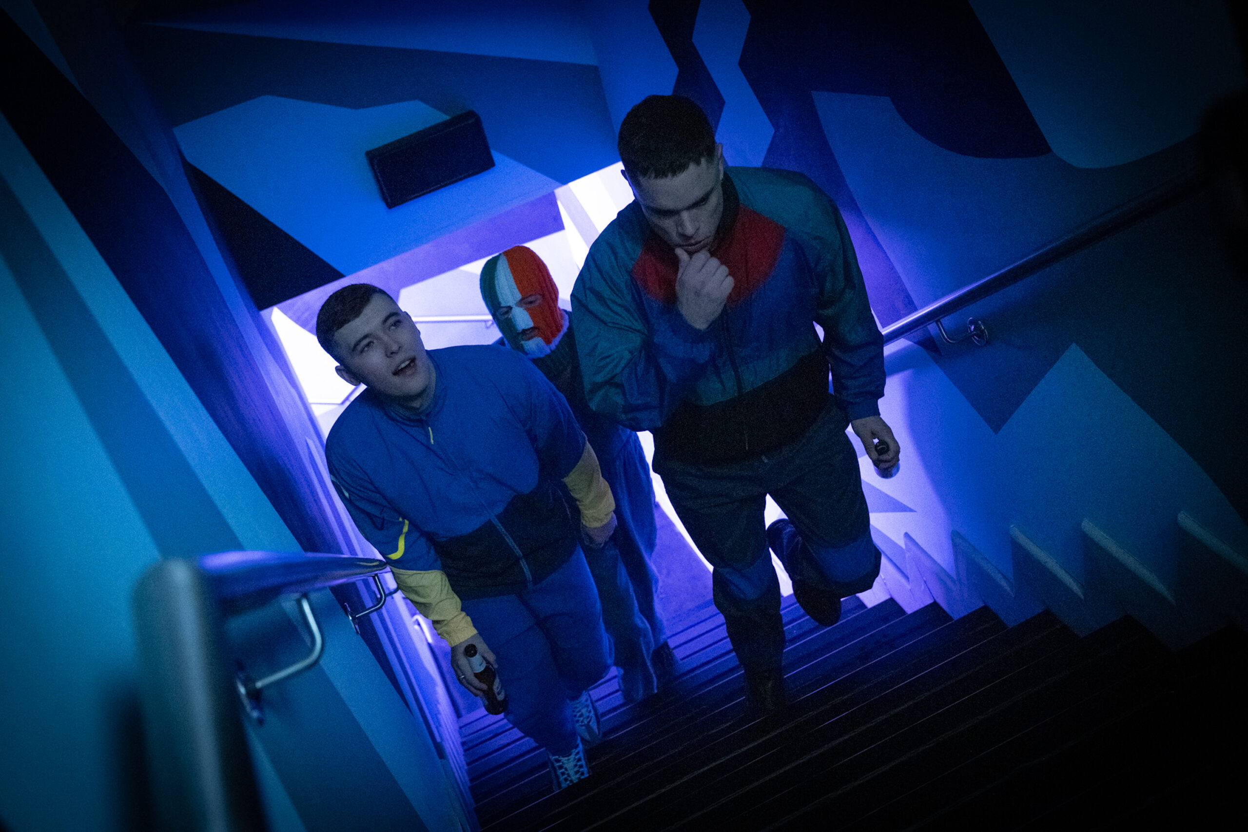 Film still from 'Kneecap' of three men walking up the stairs. One of them has an Irish flag balaclava covering his face.